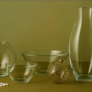 Clear glass vases, bowls, and ornaments arranged on a muted green surface, showcasing elegant craftsmanship and material clarity.