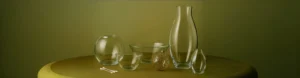 Clear glass vases, bowls, and ornaments arranged on a muted green surface, showcasing elegant craftsmanship and material clarity.