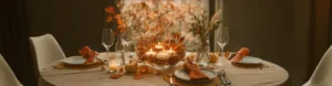 A beautifully set Thanksgiving dining table with glass vases holding orange roses and dried autumn foliage, surrounded by plates, gold cutlery, and candles, decorated with scattered fall leaves and a small pumpkin.