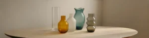 Five decorative glass vases in amber, frosted white, teal, smoky gray, and clear colors with various modern shapes on a neutral background