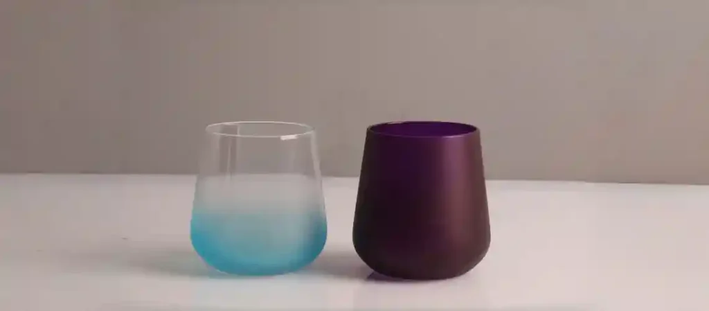 Two modern stemless wine glasses on a white surface against a neutral background: left glass with soft turquoise-to-clear ombré fade, right glass in matte deep plum purple with subtle satin finish.