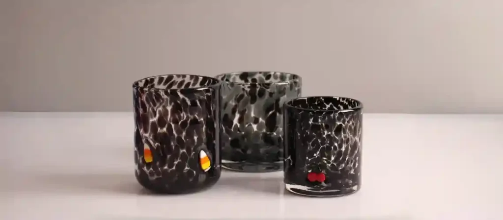 Three handmade black-and-white spotted tortoiseshell glass tumblers on a white surface against a gray background. The left glass has two small candy-corn decorations embedded in the base, the middle glass is plain smoky gray with spots, and the right glass has two tiny red cherries embedded near the bottom.