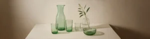 Aesthetic recycled green glassware set displayed on a neutral background, featuring bottles, tumblers, and a small bowl in soft natural light.